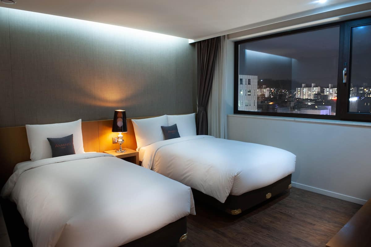 Habitación de hotel con dos camas, una mesita de noche con lámpara, ventana con vista a la ciudad y suelo de madera.