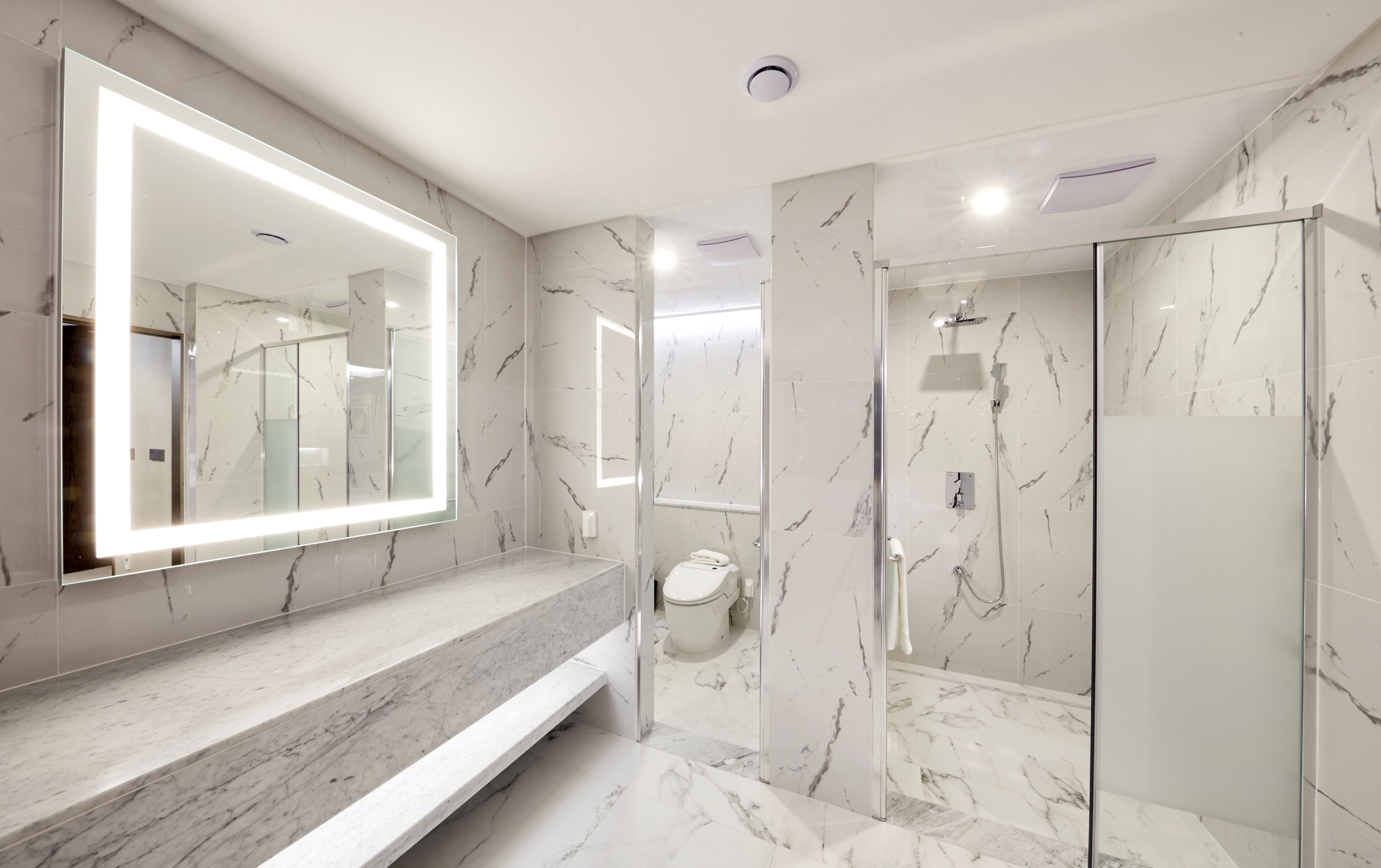 Un baño moderno con un espejo grande, encimeras de mármol, un inodoro y una ducha con mampara de cristal.