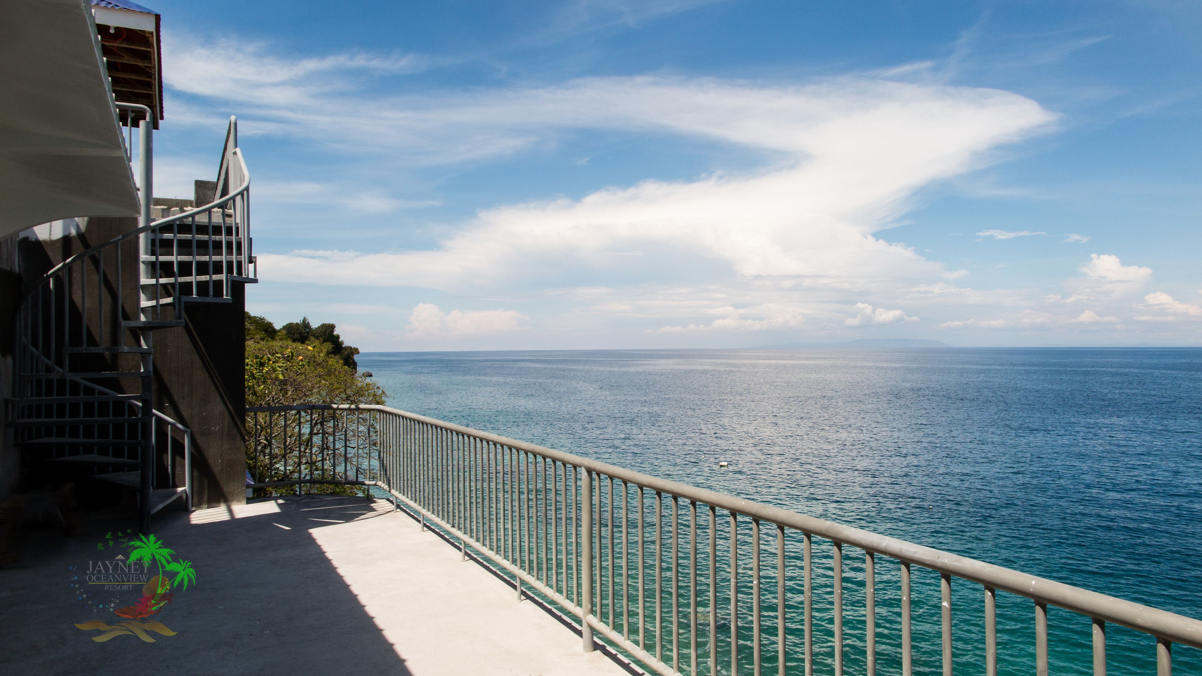 A balcony with a metal railing overlooking a body of water, a staircase on the left, and a clear sky.