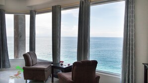 Una habitación con un ventanal grande que da al mar, dos sillones, una mesita con un florero y un mural tropical en el suelo.