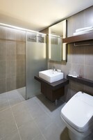 A modern bathroom with a white sink, mirror, and toilet. It features a glass shower enclosure and a dark countertop with toiletries.