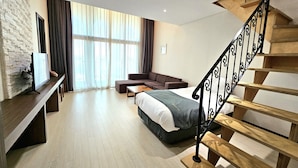 Une chambre d’hôtel moderne avec un escalier en bois, un grand lit, un canapé, un téléviseur fixé sur un mur en briques et une grande fenêtre avec des rideaux.