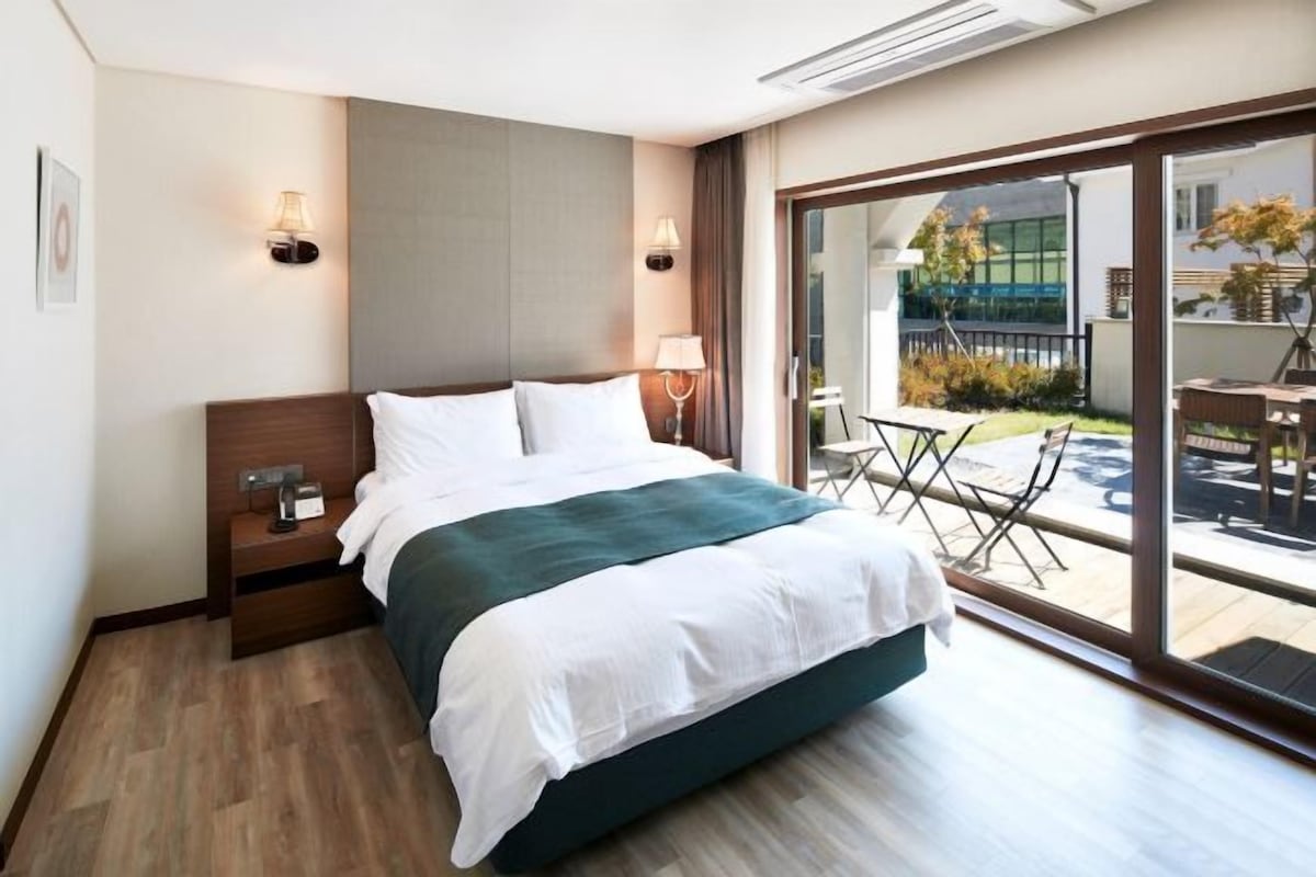 A hotel room with a bed, bedside tables, a lamp, and a view of the outdoors.