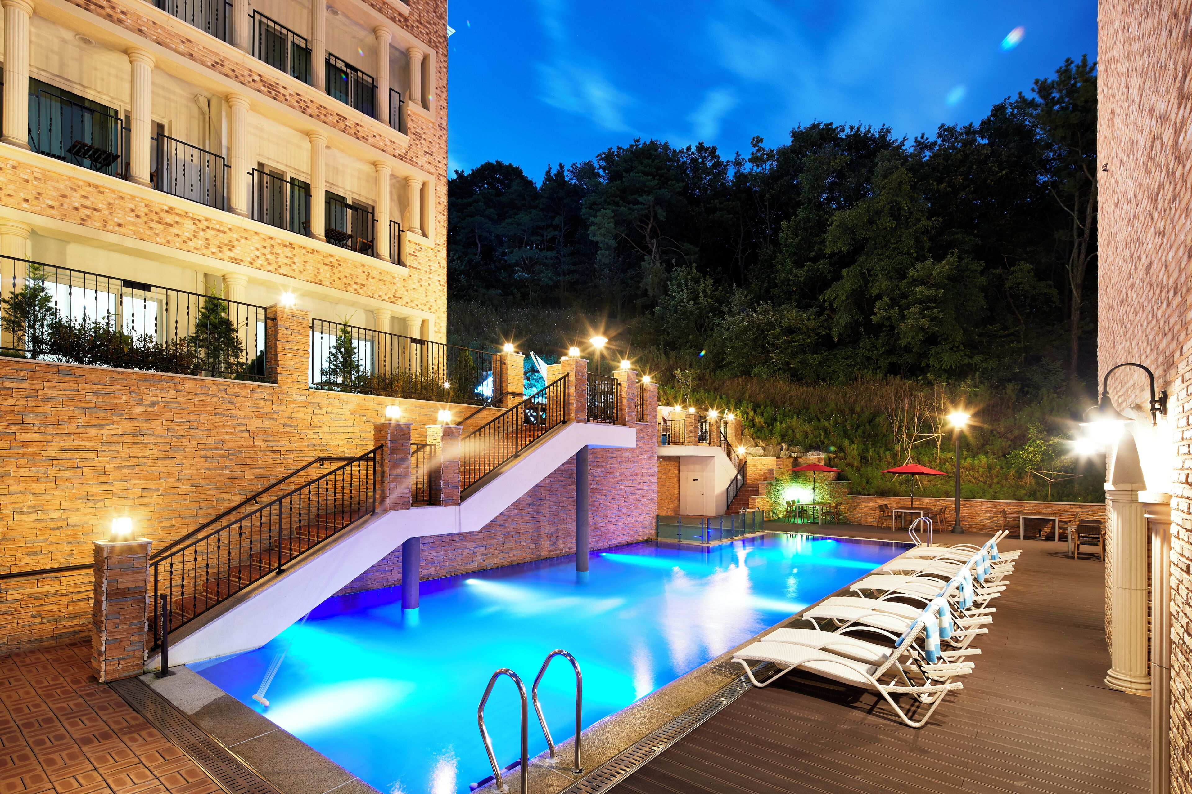 A pool area with a staircase leading to a balcony, surrounded by a brick wall and a forested hillside.