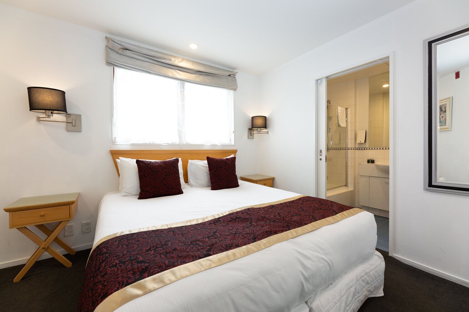 Une chambre d’hôtel bien rangée, avec un grand lit, deux tables de chevet, une fenêtre avec des rideaux et une salle de bain visible à travers une porte ouverte.