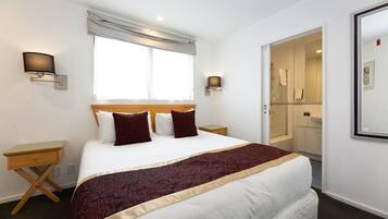 Une chambre d’hôtel bien rangée, avec un grand lit, deux tables de chevet, une fenêtre avec des rideaux et une salle de bain visible à travers une porte ouverte.