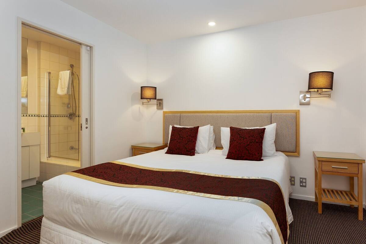 A hotel room with a bed, two bedside tables, a nightstand, a shower area, and wall-mounted lighting.