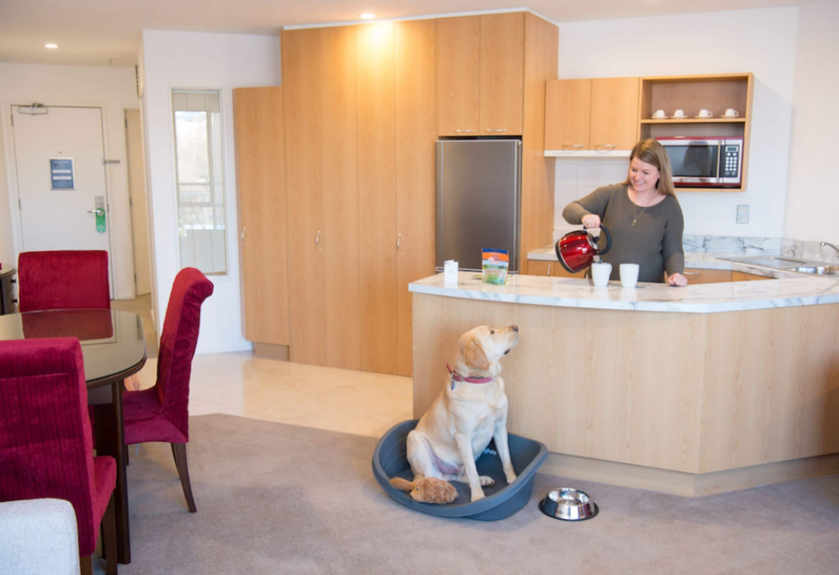 Een moderne keuken met houten kastjes, een roestvrijstalen koelkast en een magnetron. Een hond zit op een blauwe mat voor het aanrecht, en een persoon giet water in een beker.