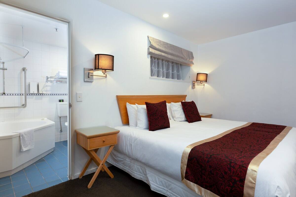 A hotel room with a large bed, two bedside lamps, a wooden nightstand, a bathroom with a shower, and a window with a curtain.
