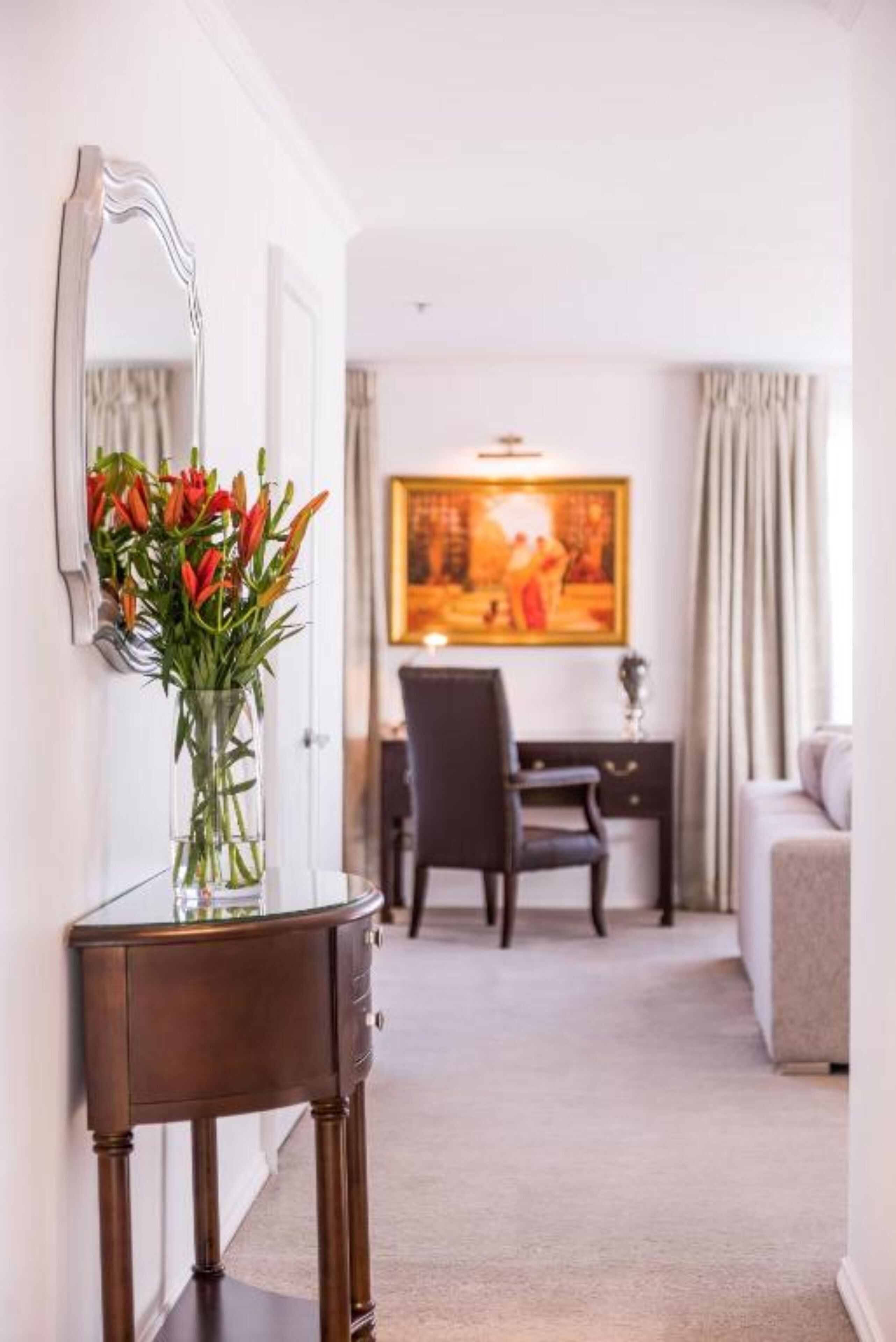 A hotel room with a wooden console table, a vase of red tulips, a large painting on the wall, and a view of the adjacent room.