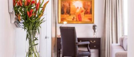 A hotel room with a wooden console table, a vase of red tulips, a large painting on the wall, and a view of the adjacent room.