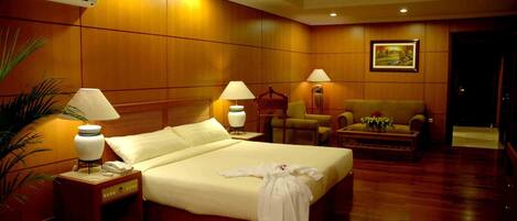 Una habitación de hotel con una cama grande, dos mesitas de noche con lámparas, una silla, una pequeña mesa con flores y un cuadro en la pared.