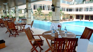 A poolside area with wooden tables and chairs, a pool, and a building with balconies in the background. - Royal Senyiur Hotel Pasuruan (Prigen)