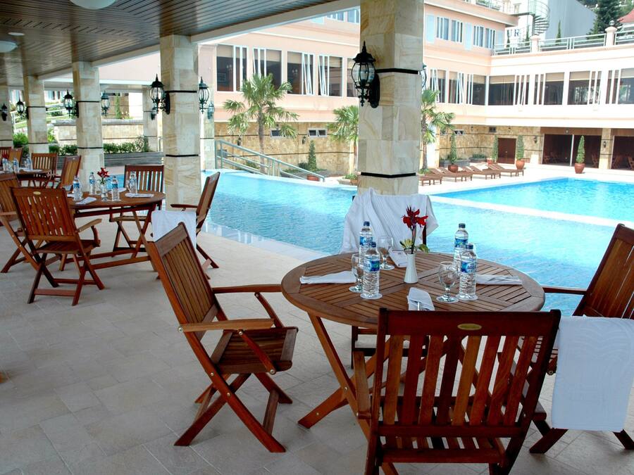A poolside area with wooden tables and chairs, a pool, and a building with balconies in the background.