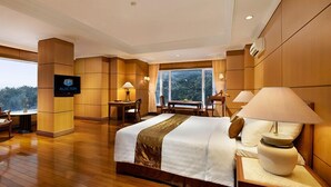 A hotel room with a large bed, a desk, a chair, a TV, and a window with a view of trees. - Royal Senyiur Hotel Pasuruan (Prigen)