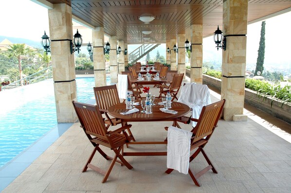 Un área de comedor al aire libre techada con vista a la piscina, que incluye una mesa redonda con sillas y mantel blanco.