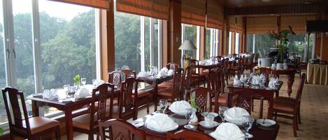 Un comedor con varias mesas dispuestas para el servicio, amplias ventanas con vistas a árboles y un techo con vigas de madera.