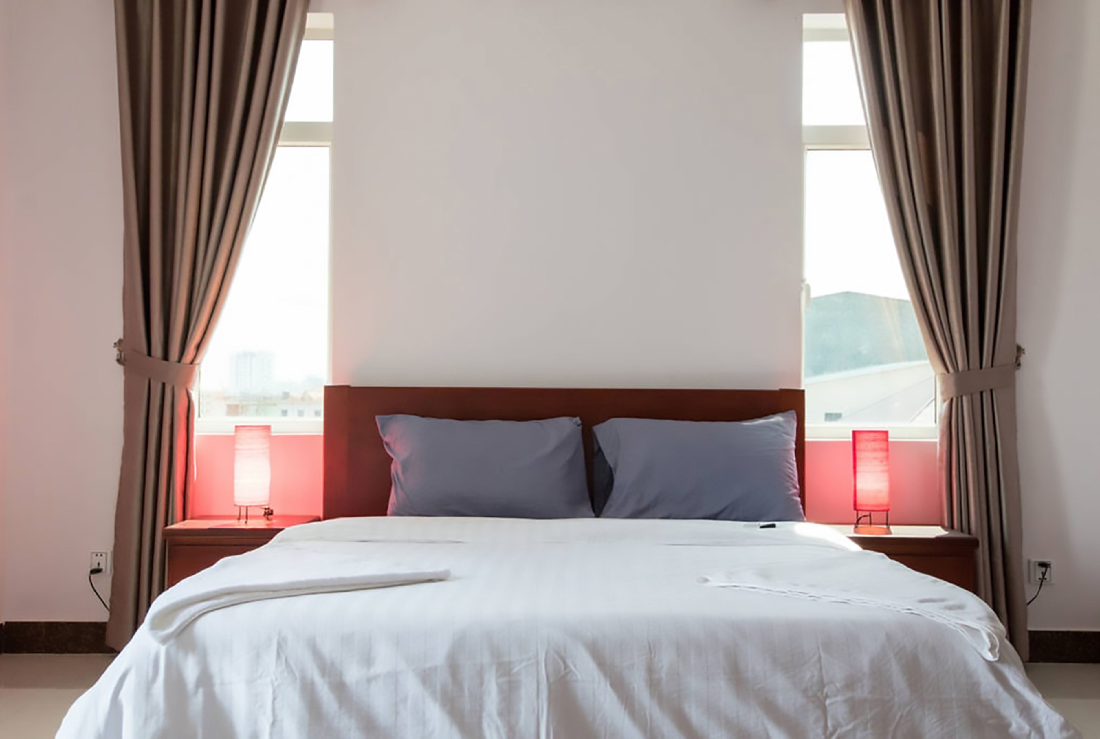 Een hotelkamer met een groot bed, twee kussens, een nachtkastje, een raam met gordijnen en een rode lamp.