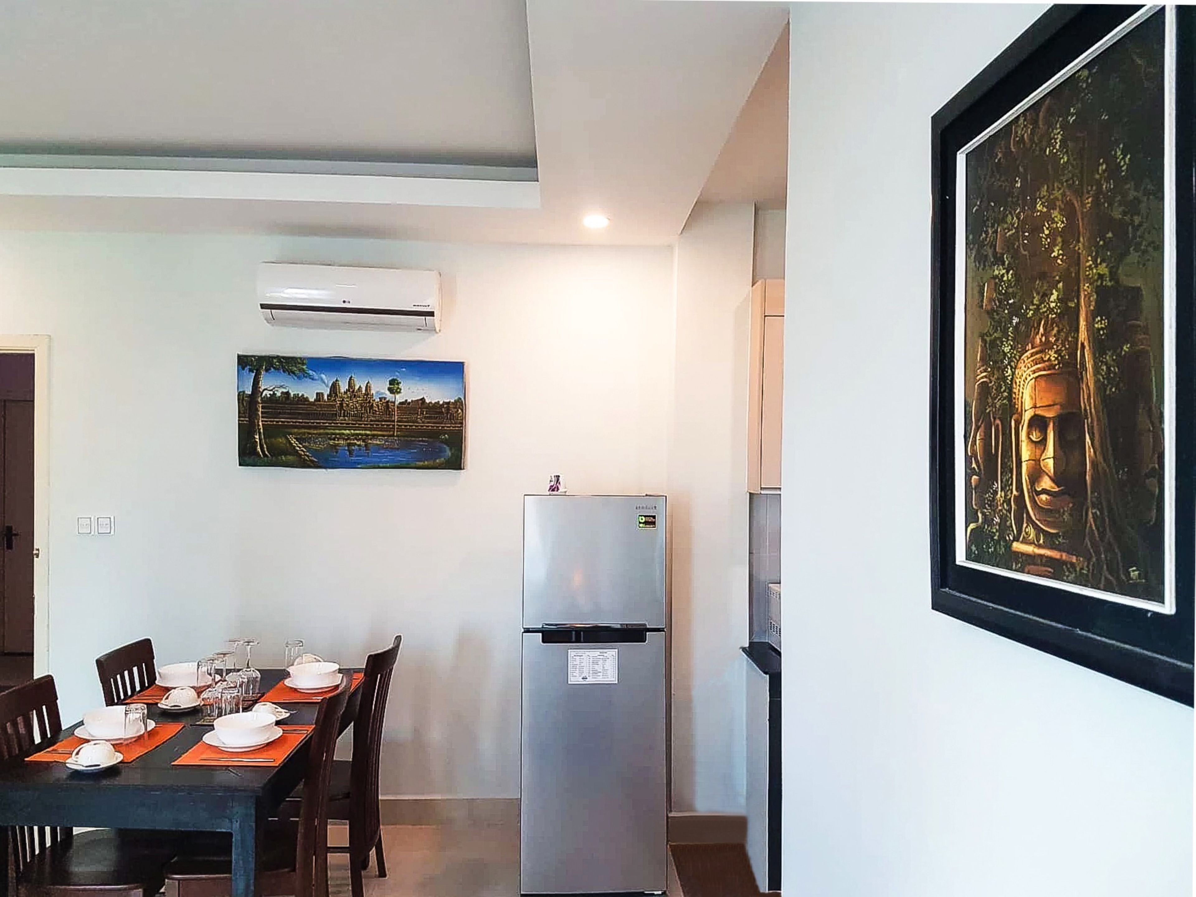 A dining area with a table set for four, a refrigerator, and a Buddha statue on the wall.