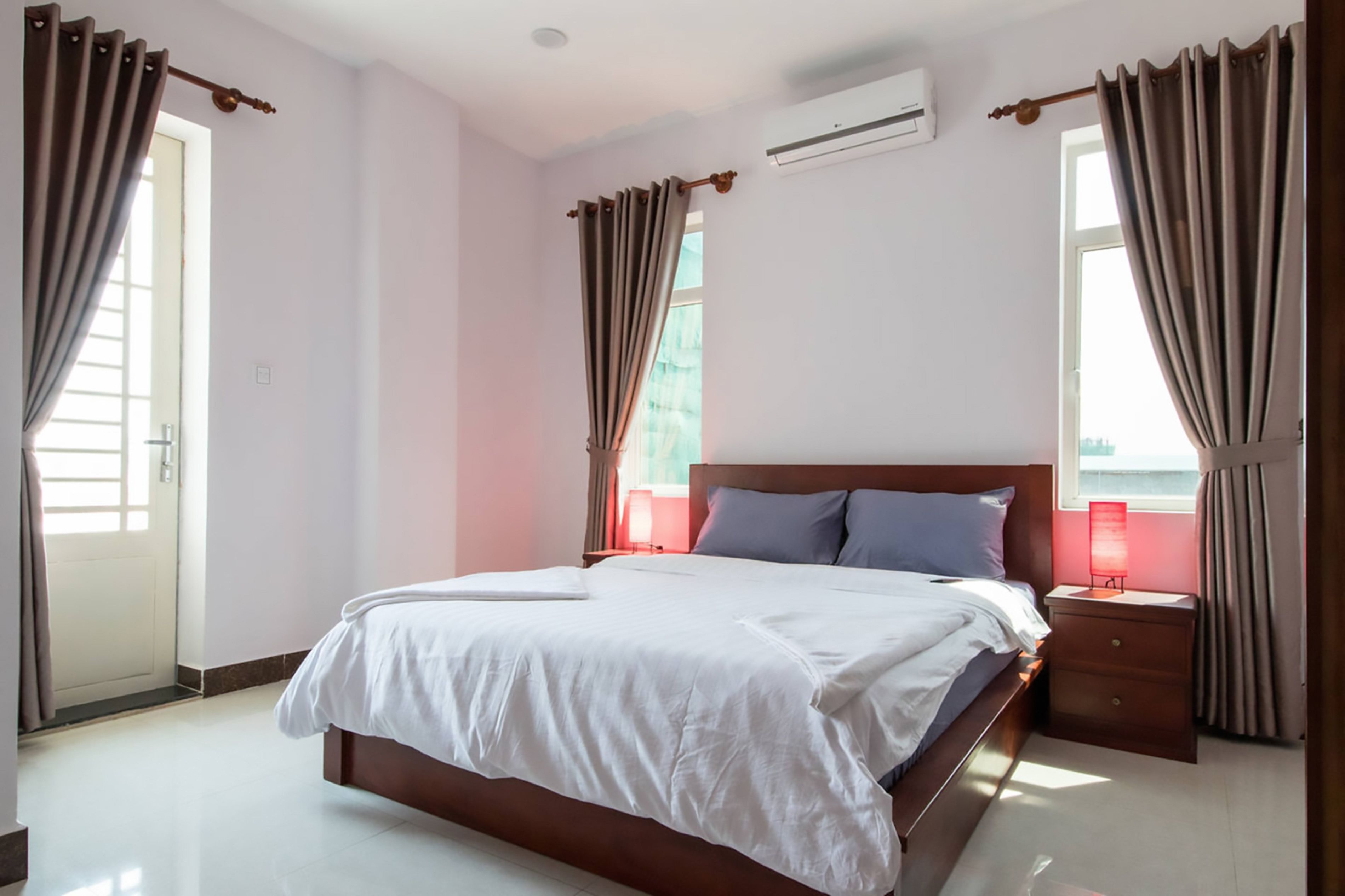 Ein Hotelzimmer mit einem großen Bett, einem Nachttisch, einer roten Lampe und einem Fenster mit Vorhängen.