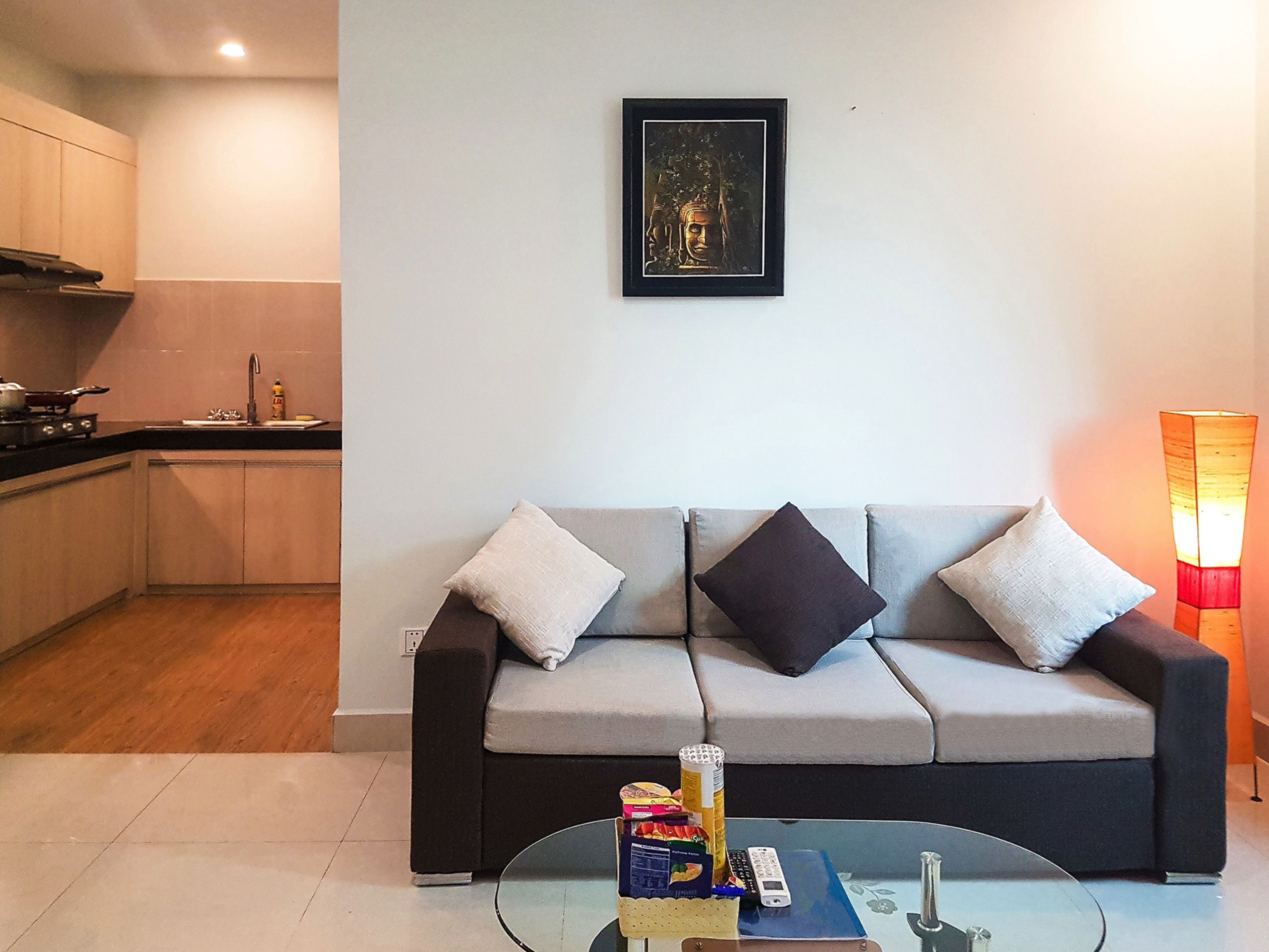 A modern living room with a grey sofa, a glass coffee table, and a kitchen area in the background.