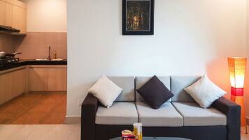 A modern living room with a grey sofa, a glass coffee table, and a kitchen area in the background.