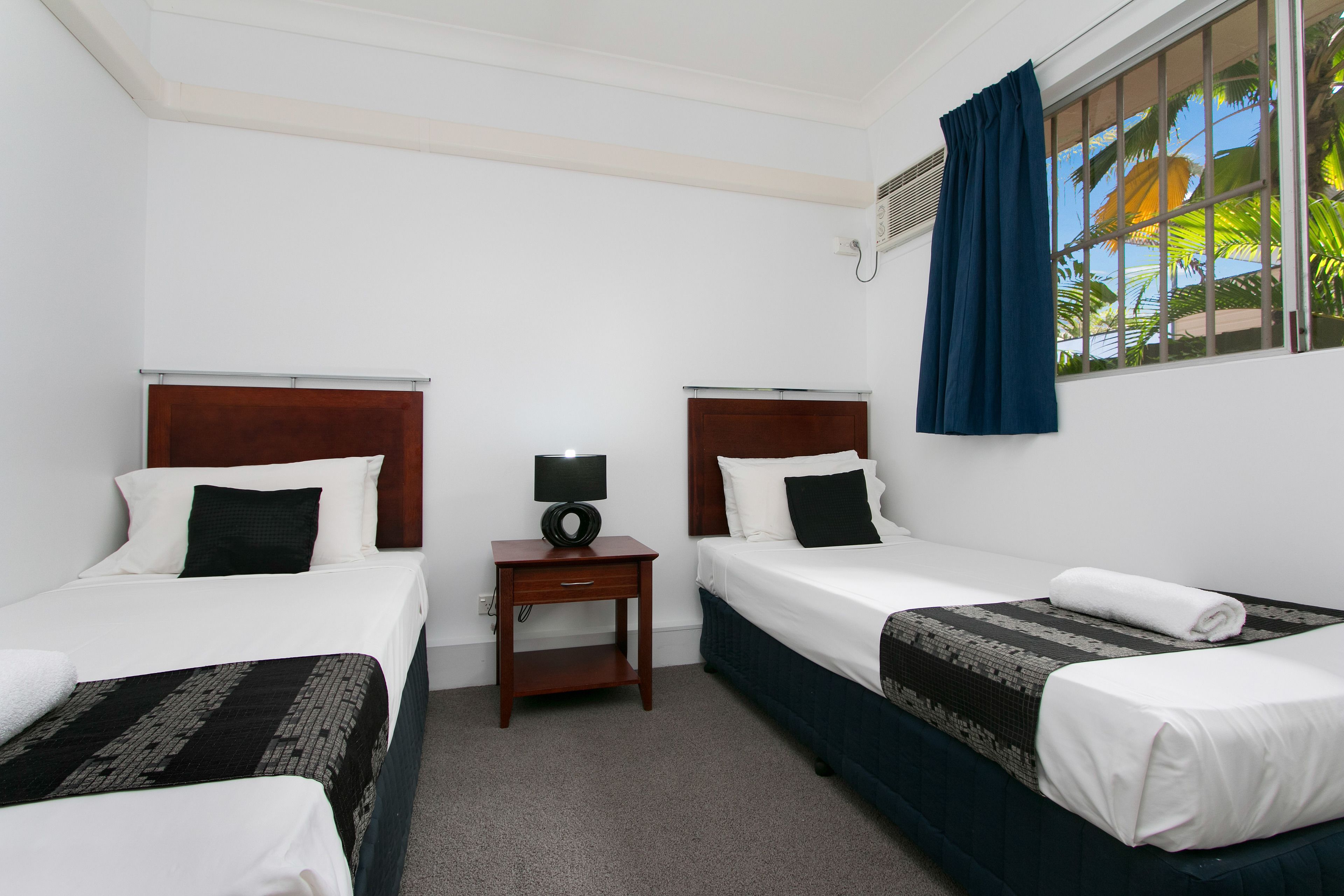 Una habitación de hotel con dos camas individuales, una mesita de noche de madera, una lámpara y una ventana con cortinas azules.