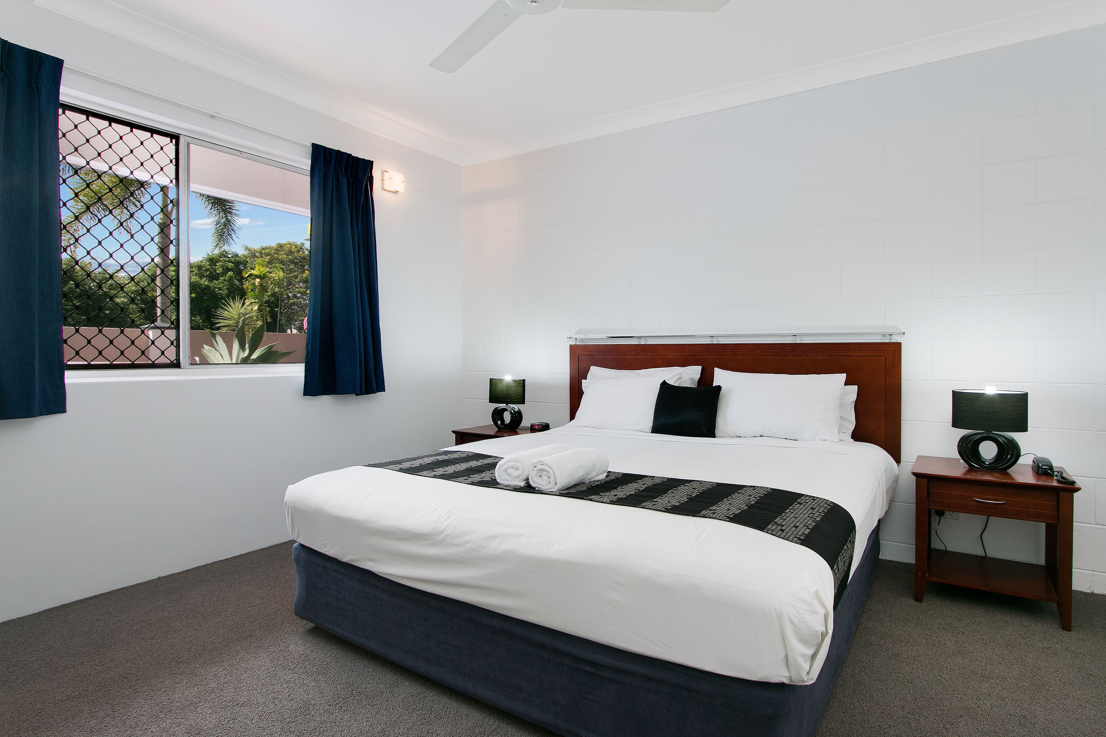 A neatly made bed with white linens and a dark blue accent, two bedside tables with lamps, a wooden headboard, and a window with blue curtains.