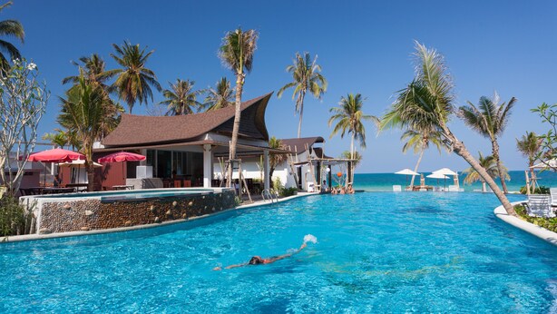 Ein Swimmingpool mit einer Person, umgeben von Palmen und einem Gebäude mit einem geneigten Dach.