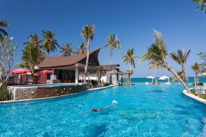 Piscina com uma pessoa nadando, cercada por palmeiras e um edifício com telhado inclinado.