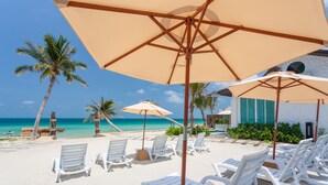 Eine Strandlounge mit weißen Liegestühlen und beigen Sonnenschirmen, Palmen und freiem Blick auf das Meer.
