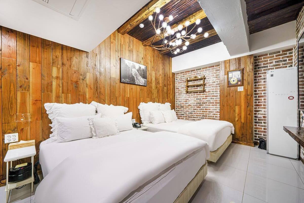 Quarto moderno com duas camas, painéis de madeira, parede com detalhes em tijolo e luminária no teto.