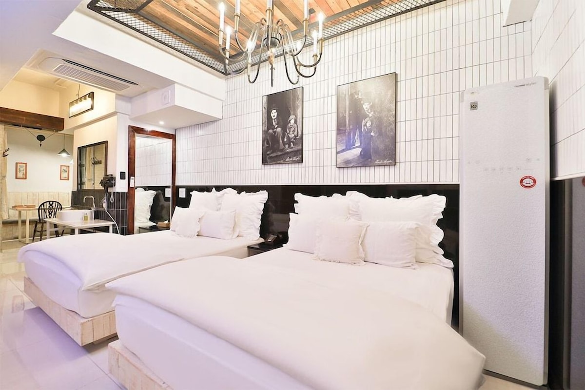 Habitación de hotel con dos camas, un candelabro, un refrigerador y cuadros en la pared.