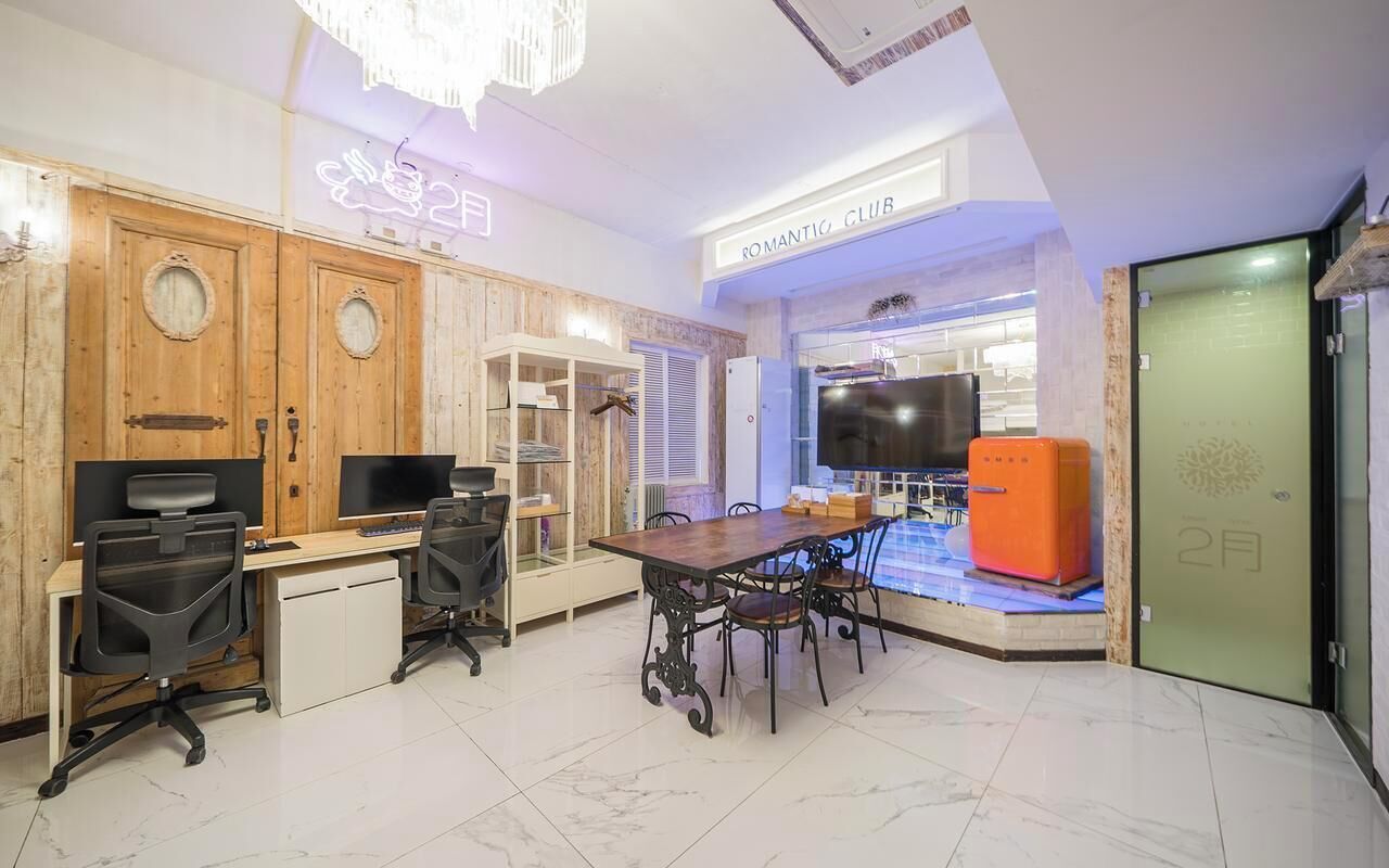 Una habitación de hotel moderna con un escritorio de madera, dos sillas de oficina, una mesa de comedor con sillas, un televisor y un letrero de neón.