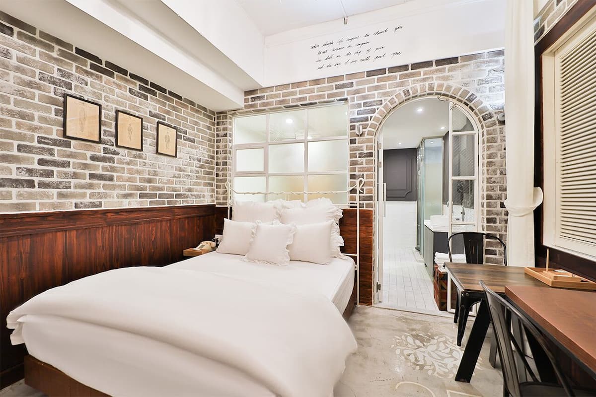 Quarto com parede de tijolos, cabeceira de madeira, cama grande, escrivaninha com cadeira e espelho.