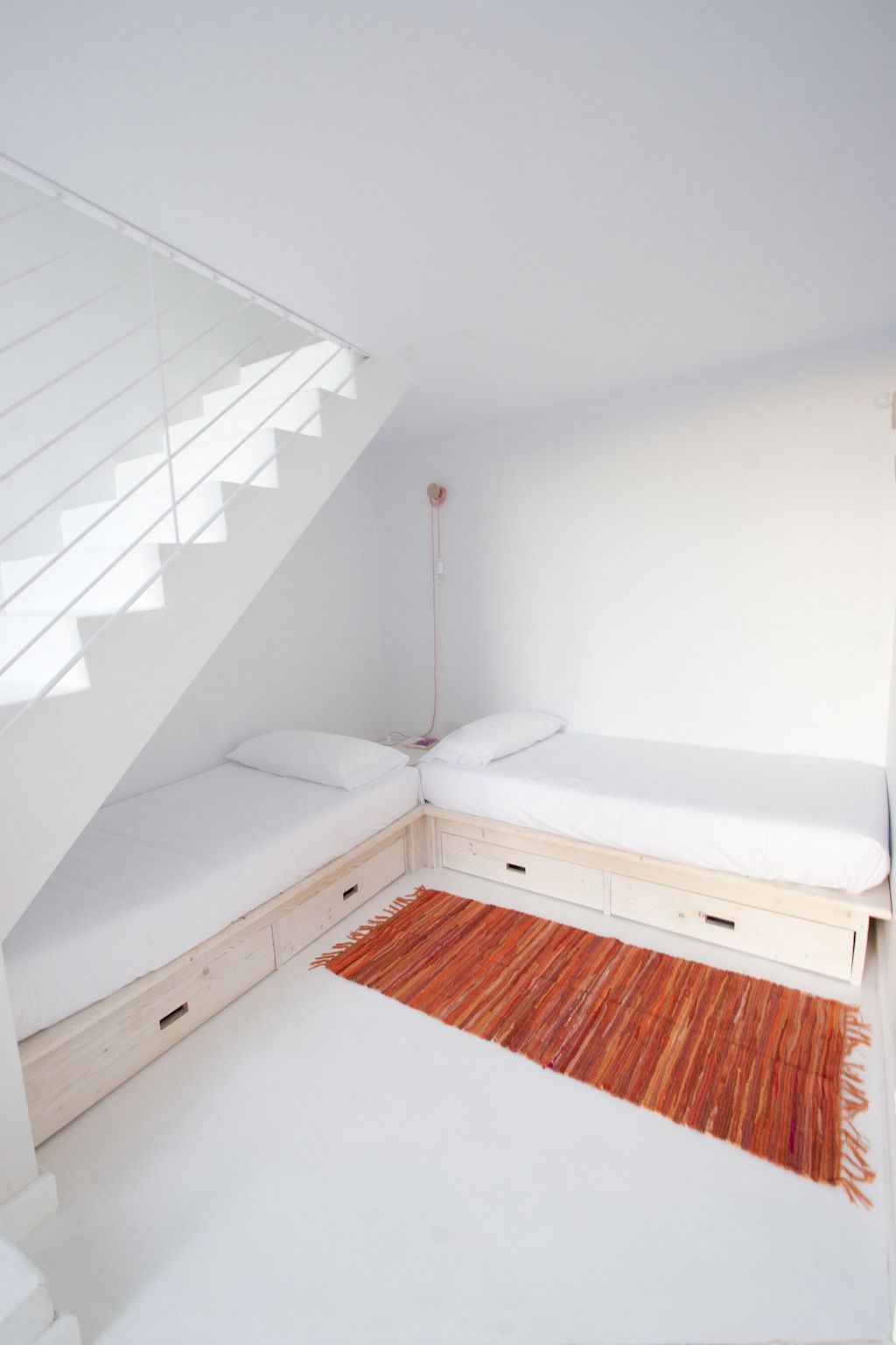 Un dormitorio moderno y minimalista con techo inclinado, una cama empotrada con cajones de almacenamiento y una alfombra roja tejida.
