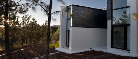 A modern building with large windows and a stone facade, surrounded by a forested area.