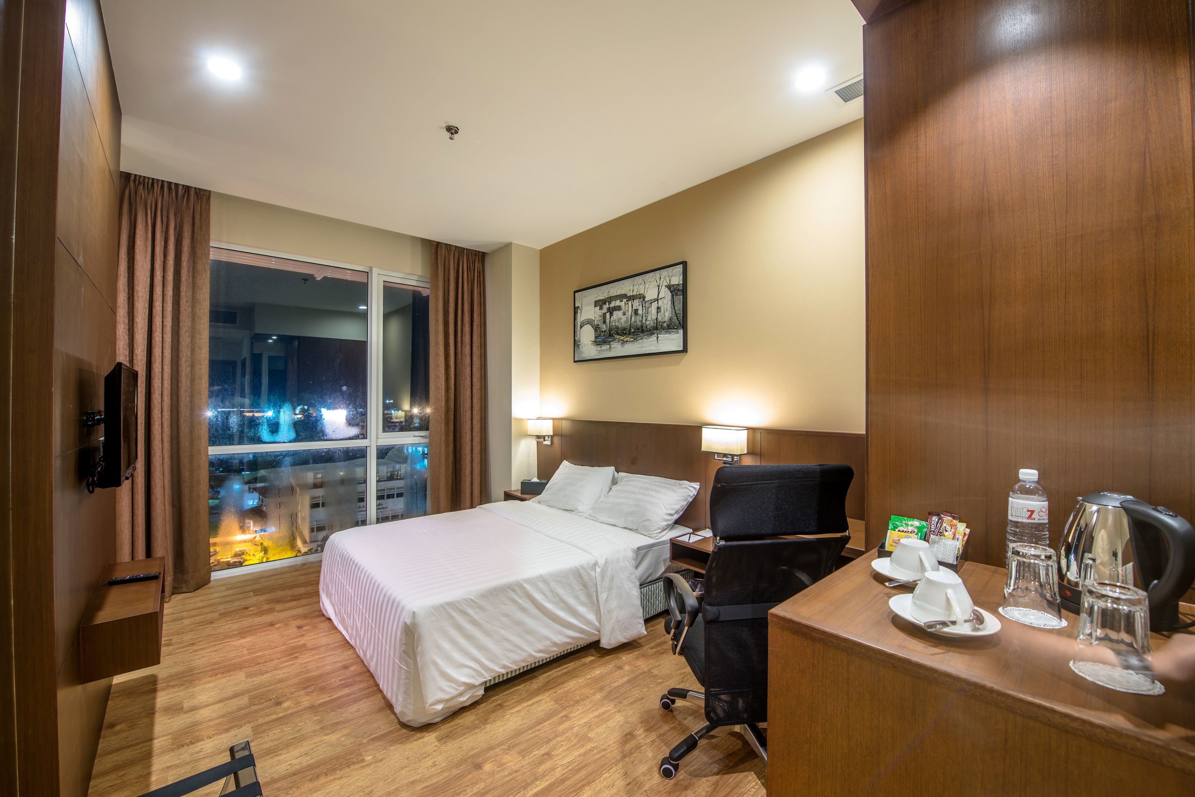 A hotel room with a bed, desk, chair, and a window with a view of a cityscape at night.