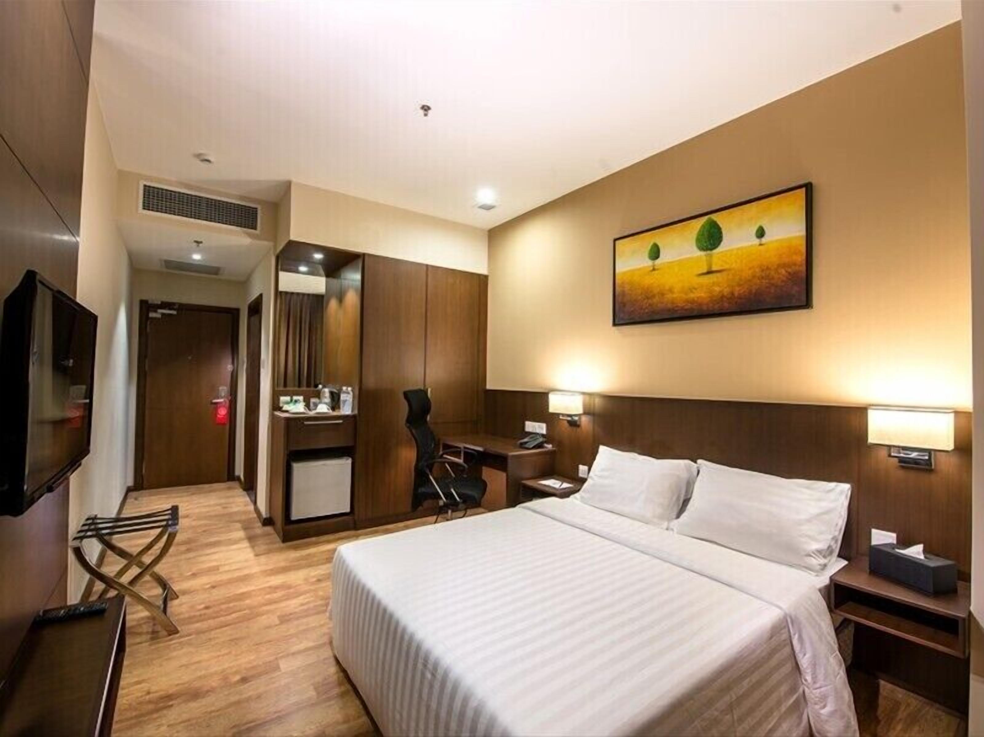 A hotel room with a bed, desk, chair, TV, and artwork on the wall.