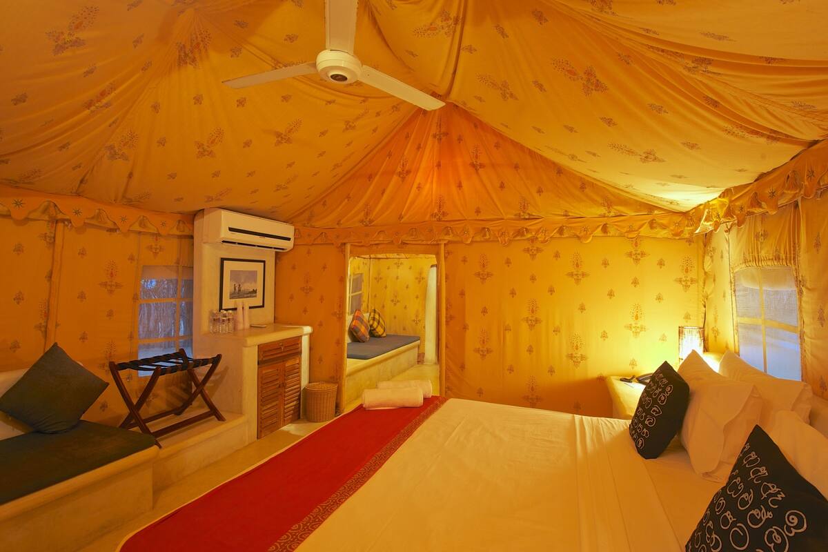 Quarto estilo tenda com cama, travesseiros, um tapete vermelho, uma mesinha e um aparelho de ar condicionado.