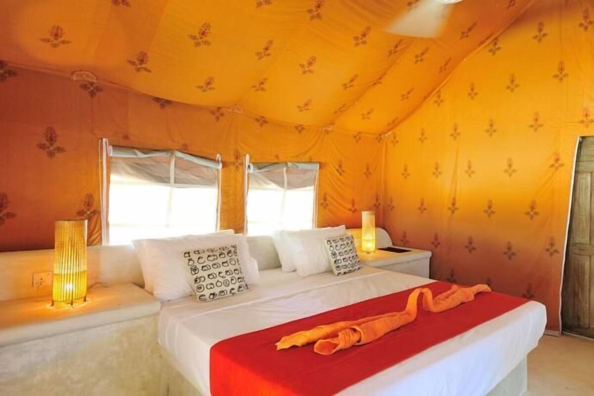 Quarto com cama, duas mesinhas de cabeceira, um criado-mudo, uma luminária e uma porta.
