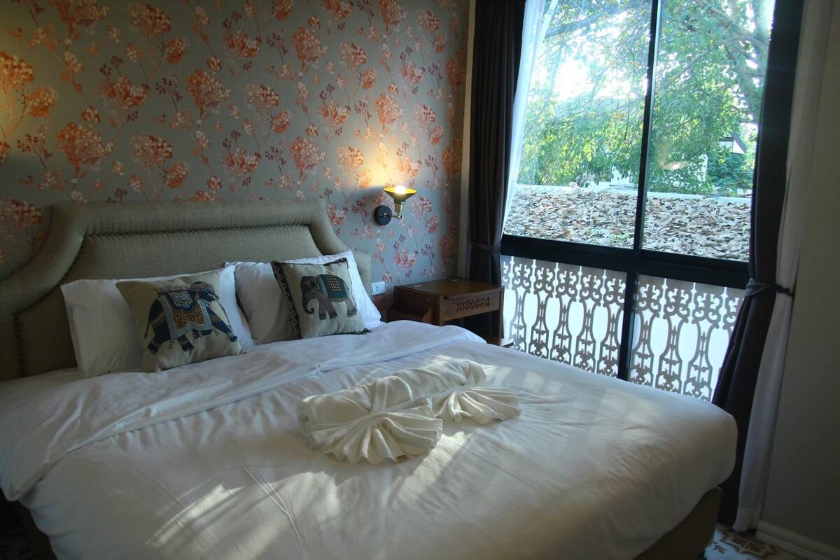 A neatly made bed with decorative pillows, a wall-mounted light, and a window with a view of trees.