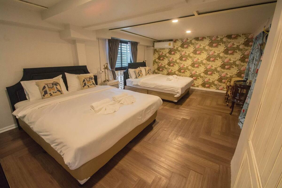 A hotel room with two beds, wooden flooring, floral wallpaper, and an air conditioning unit.