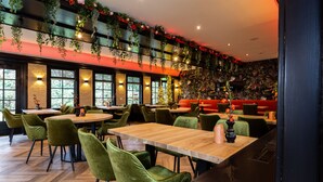 Un restaurante con mesas de madera y sillas de terciopelo verde, rodeado de grandes ventanales y una pared decorada con plantas y flores.