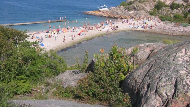 Una playa con aguas cristalinas, terreno rocoso y un barco a lo lejos.