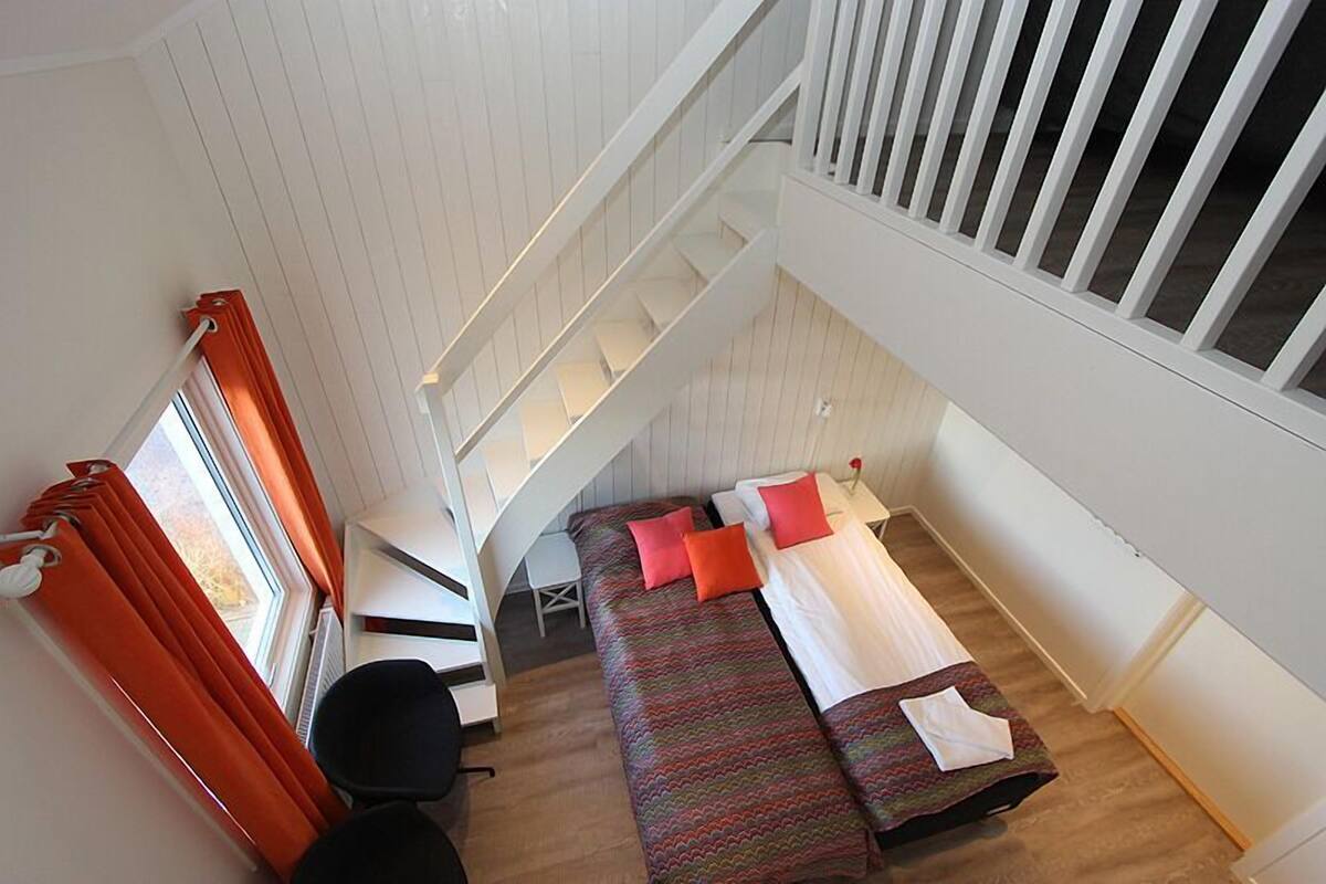 A compact room with a bed, a desk, and a chair. There is a window with curtains and a view of the outdoors.
