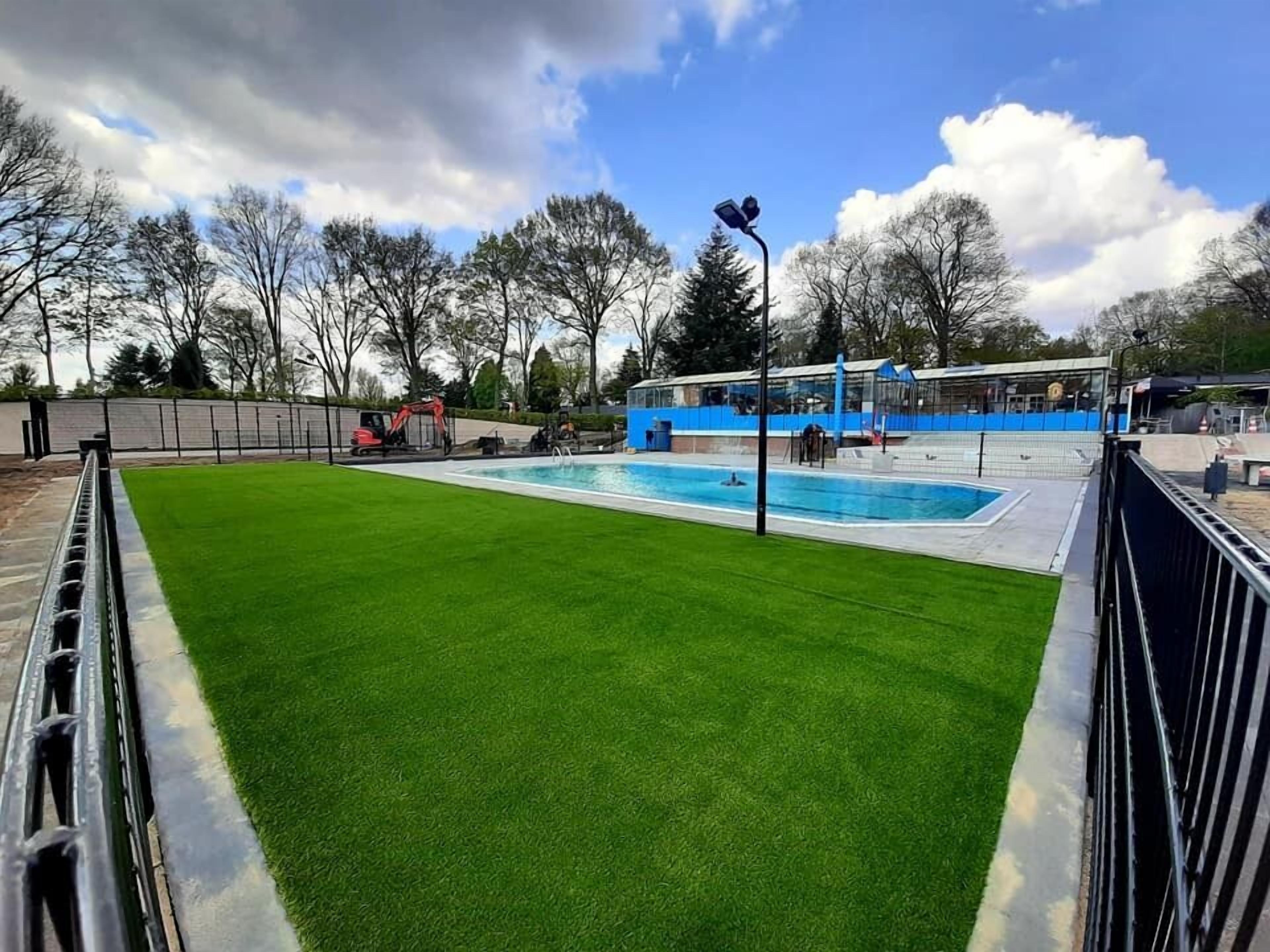 A swimming pool with a surrounding grassy area, a blue building, and a fenced playground.