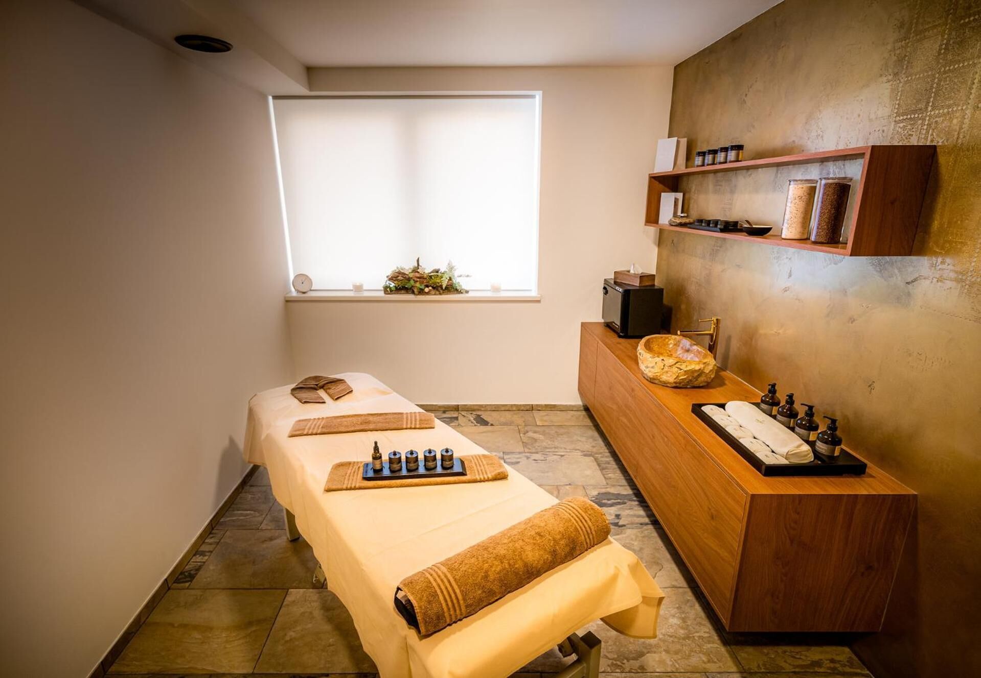 A massage room with a wooden massage table, a wooden reception desk, a window with a view, and shelves with jars and a clock.
