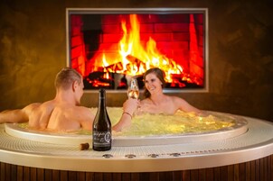 Dos personas en un jacuzzi con una botella de cerveza y un corcho, frente a una chimenea.