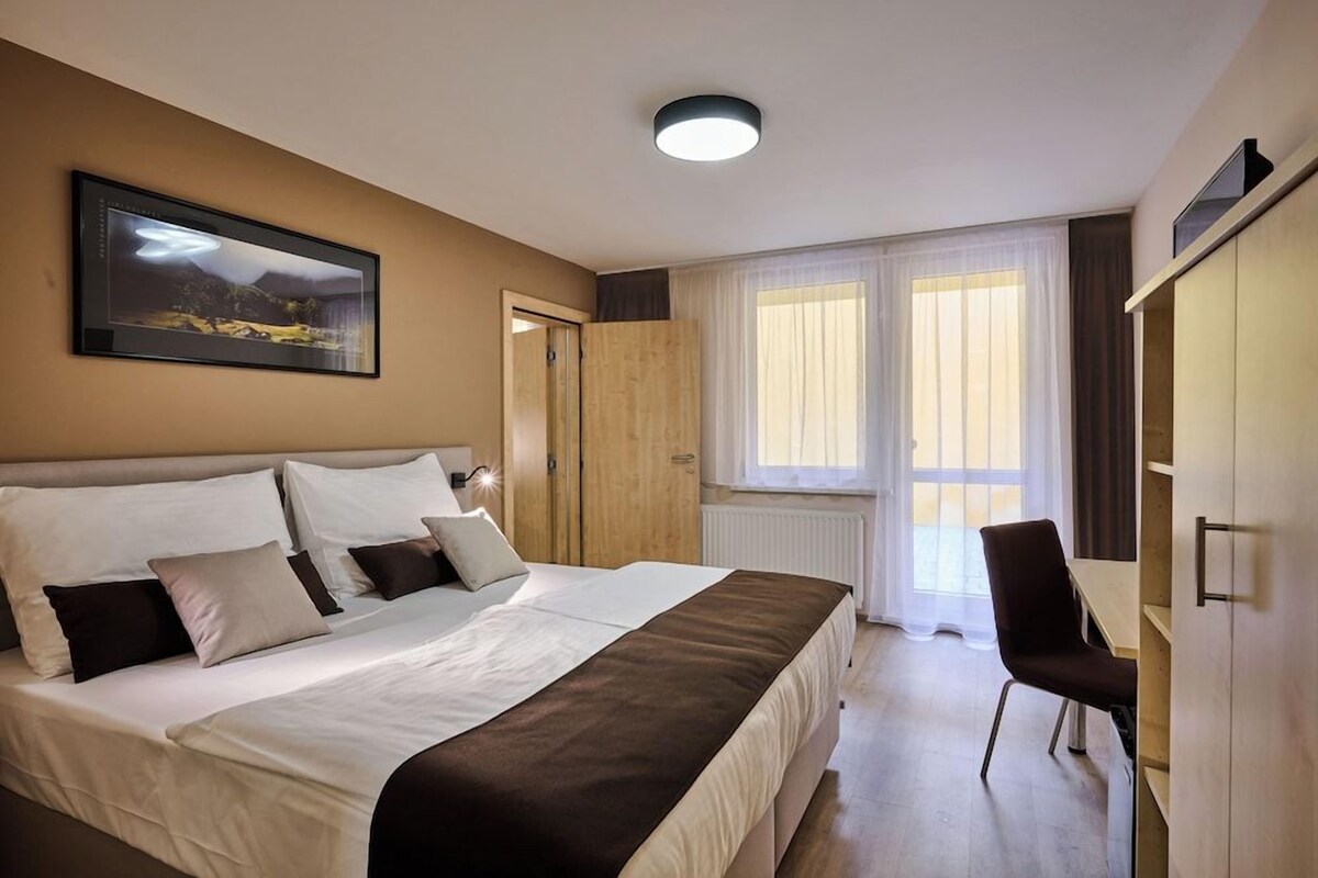 Una habitación de hotel con una cama, un escritorio, una silla, un ventanal con cortinas y un cuadro en la pared.